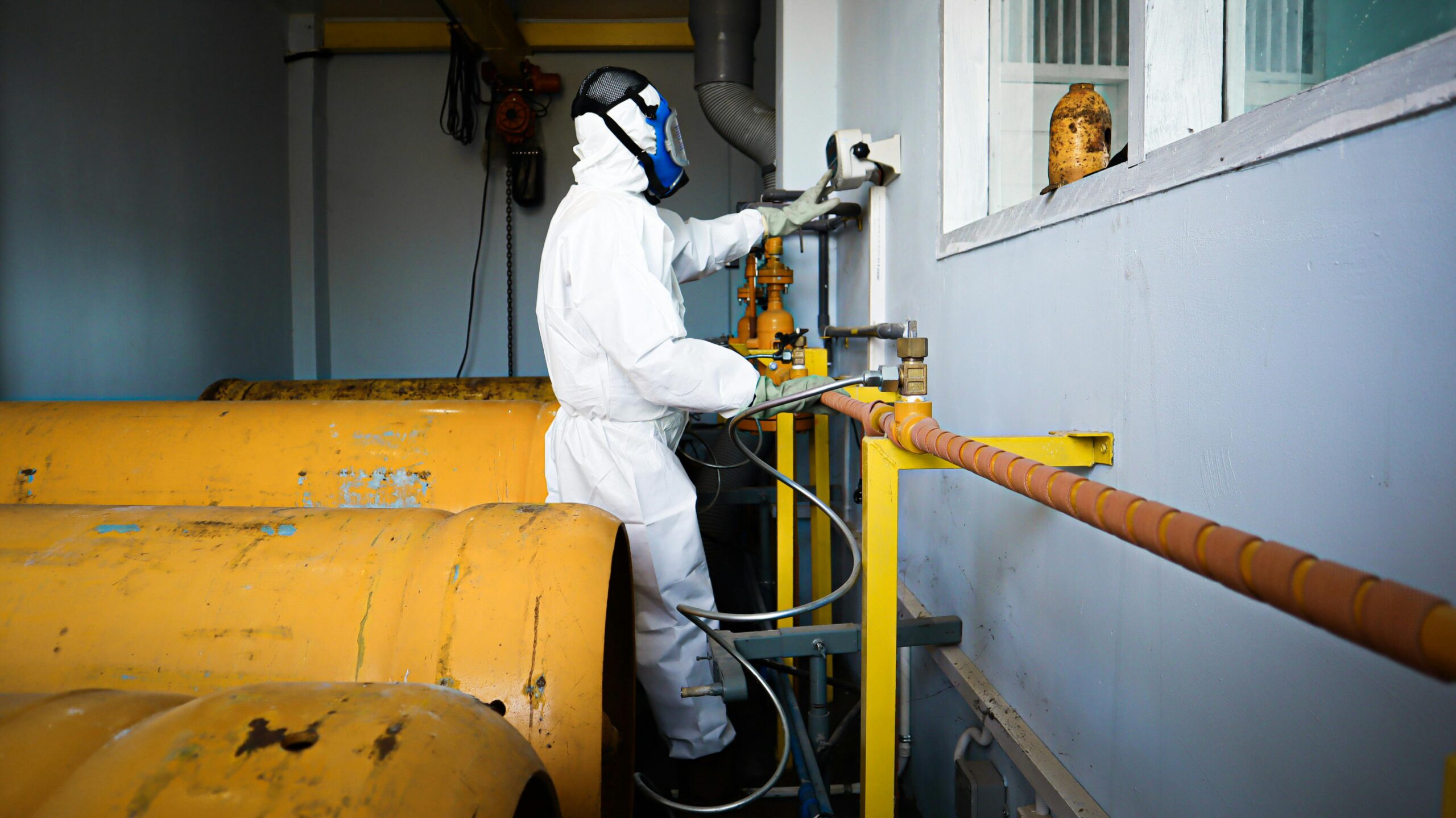 Worker in full protective suit and gas mask handles industrial pipes indoors in Indonesia.